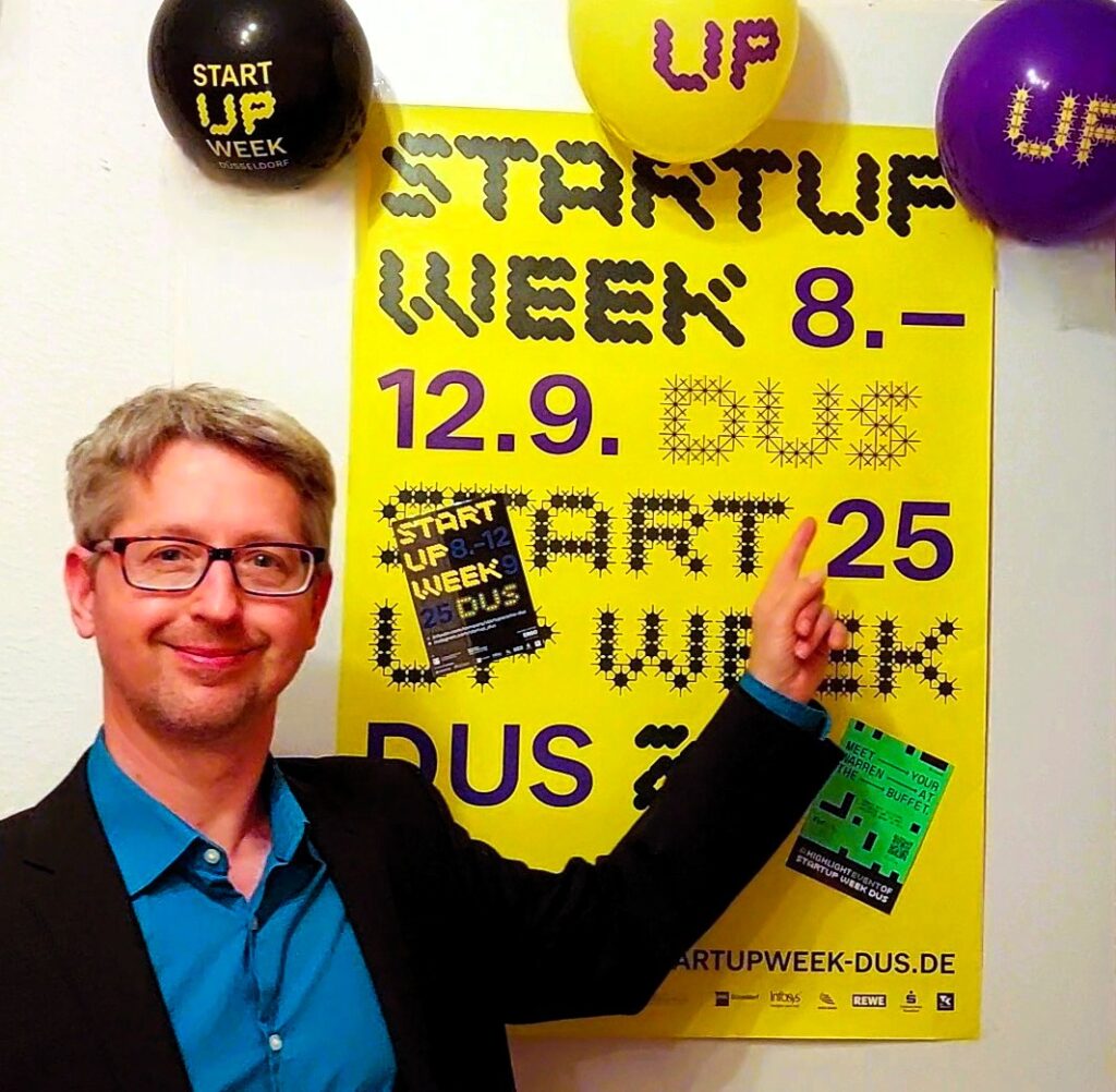 Beschreibung Headerfoto: Stefan Klemens zeigt auf ein gelbes Poster der Startup Woche Düsseldorf 2025.