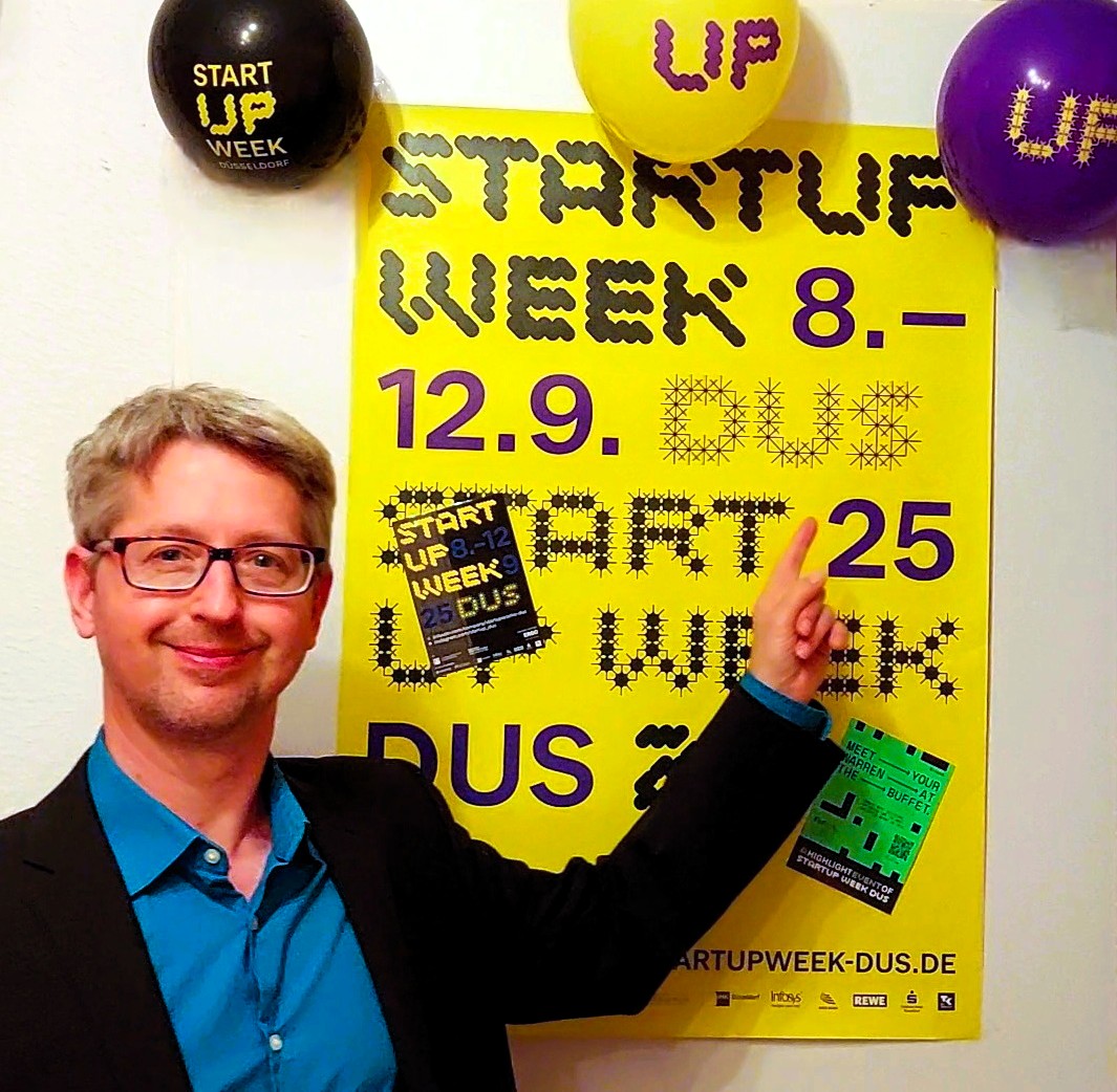 Beschreibung Headerfoto: Stefan Klemens zeigt auf ein gelbes Poster der Startup Woche Düsseldorf 2025.